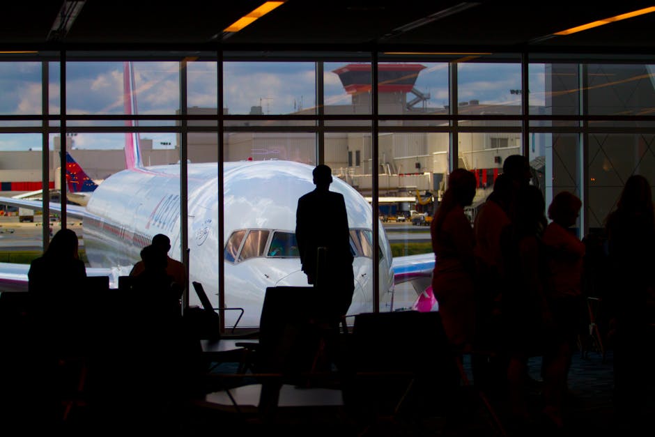A traveler sitting by an airport gate window checking their phone, with a plane visible on the tarmac outside