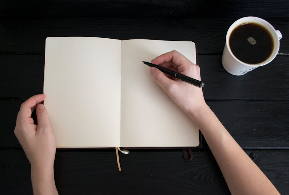 A hand writing in an open notebook on a light-colored table in warm afternoon light