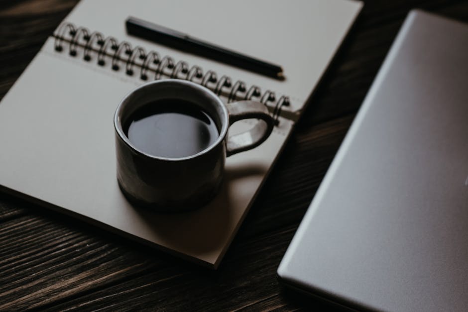 A minimal wooden desk with a small notebook, pen, and espresso in soft natural light
