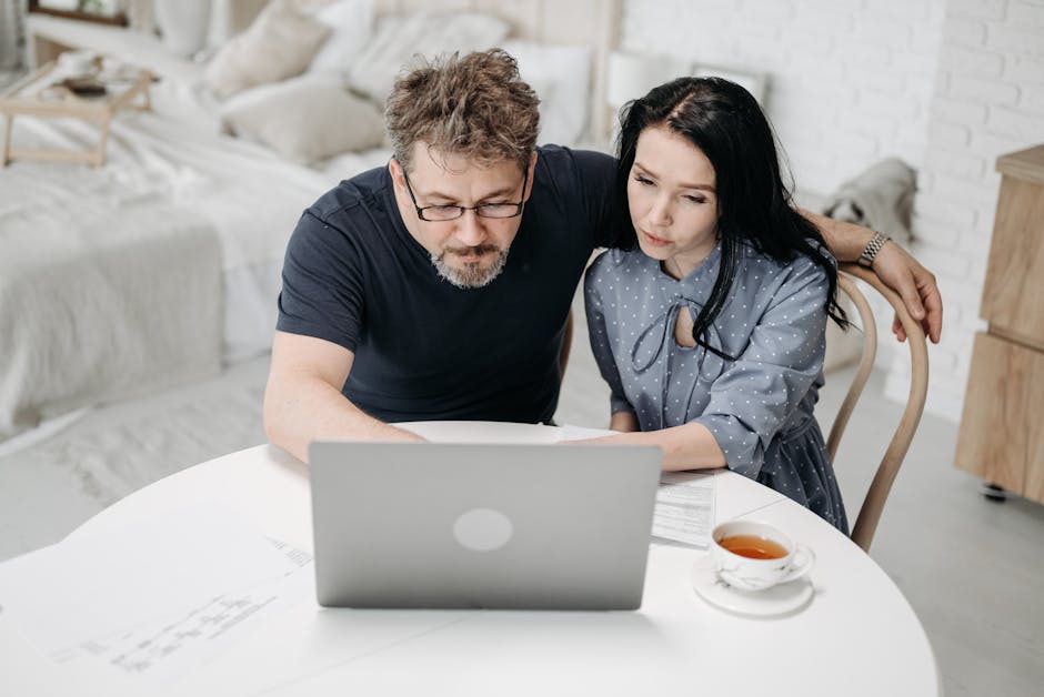 Two people sitting together at a kitchen table looking at a laptop with papers and a coffee nearby in warm afternoon light