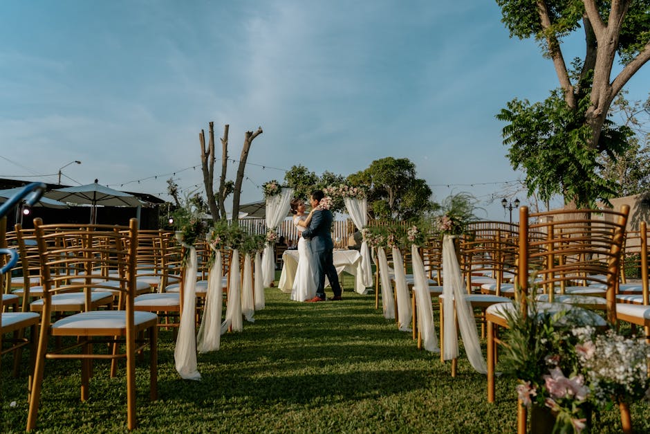 An outdoor wedding ceremony with guests seated on wooden chairs in soft golden hour sunlight with minimal decoration