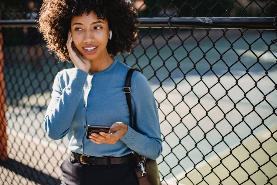 A person wearing earbuds walking on a quiet city street, glancing at their phone in natural light