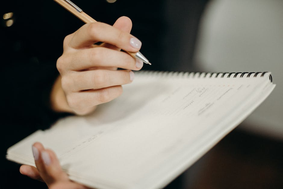 A person writing in a short notebook list with a pencil on a clean white table