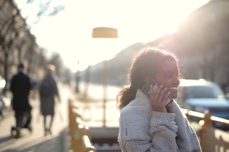 A person walking outdoors in warm sunlight, talking naturally on their phone with a relaxed expression